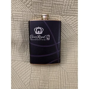 May include: Black flask with gold trim and a gold cap. The flask features the Crown Royal logo and the NBA logo, with a basketball court design. The text reads "Crown Royal Proud Partner of the NBA."