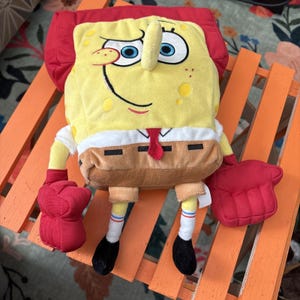 May include: A SpongeBob SquarePants soft toy. The yellow character has a red hat, red gloves, and black shoes. He wears a white shirt, brown trousers, and a red tie. The toy is sitting on an orange wooden chair.