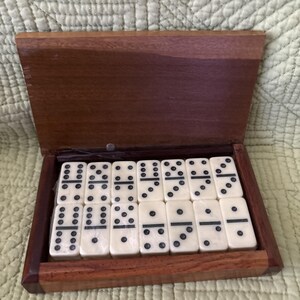 May include: A rectangular wooden box with a hinged lid containing a set of fourteen off-white dominoes arranged in two rows. Each domino features black pips and a central horizontal line, showcasing various numerical combinations in a classic game set.
