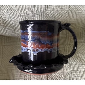 Op de afbeelding: Een donkergekleurde keramische mok met een glanzende afwerking, voorzien van een horizontale band van gemengd blauw, paars en koperkleurig glazuur. De mok staat op een bijpassende schotel met een gegolfde rand en een gebogen handvat.