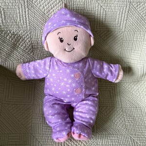 May include: A plush baby doll wearing a lavender onesie and matching cap, both adorned with white snowflake patterns. The doll has a light skin tone, rosy cheeks, and a gentle smile. The outfit is fastened with two pink buttons.