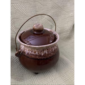 Può includere: Una pentola in ceramica marrone con coperchio e manico in metallo. La pentola ha una finitura maculata e smaltata con un bordo più chiaro. Il coperchio ha un piccolo pomello arrotondato. La pentola viene probabilmente utilizzata per cucinare o servire.