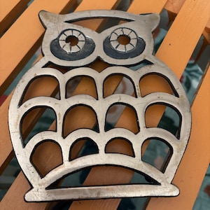 May include: Silver-colored metal owl-shaped trivet. The trivet features a cutout design with a series of arched shapes and two large eye sockets. The owl's eyes have a detailed, circular pattern. This decorative item is designed to protect surfaces from hot items.