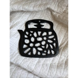 Puede incluir: Salvamanteles negro de hierro fundido con forma de tetera, con asa y boquilla. El salvamanteles presenta un diseño calado decorativo con varios recortes ovalados y circulares. Está diseñado para proteger las superficies de los utensilios de cocina calientes.