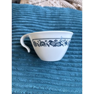 May include: White ceramic teacup with a curved handle and a blue floral pattern around the rim. The cup is set against a blue textured background. The teacup is a classic design.