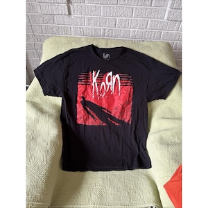 May include: Black t-shirt featuring the band name "Korn" in white, stylized text over a red and black graphic. The shirt has a crew neck and short sleeves. The band's logo is also printed on the top of the shirt.