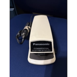 May include: Cream-colored Panasonic commercial electric stapler with a black base and a black power cord. The stapler has the brand name and product description printed in black on a black rectangle.