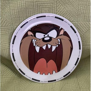 Puede incluir: Un plato redondo de color blanco que presenta un gráfico central grande del personaje Taz, el Demonio de Tasmania de Looney Tunes, con la boca muy abierta y dientes afilados. El borde exterior está decorado con el texto negro Looney Tunes.