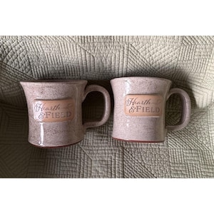 Op de afbeelding: Twee bespikkelde keramische mokken met handvatten. Elke mok heeft een rechthoekig etiket met de woorden "Hearth & FIELD" in een scriptlettertype. De mokken zijn lichtbruin van kleur en staan op een gewatteerd groen oppervlak.