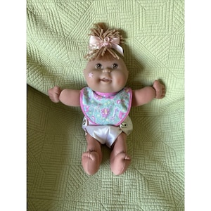 May include: A doll with brown yarn hair tied with a pink ribbon, featuring a small star decal on the cheek. The doll wears a light blue bib with a colorful animal print pattern and a white diaper with silver snaps, showing a soft, rounded body.