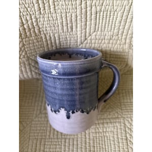 May include: A ceramic mug featuring a two-tone glaze with a deep blue upper section that drips down into a lighter, off-white base. The mug has a cylindrical body with subtle horizontal ridges and a curved handle on the right side.