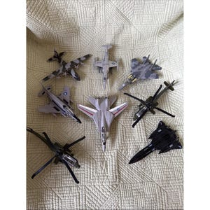 Può includere: Una collezione di aerei ed elicotteri giocattolo in vari colori e design. L'assortimento comprende caccia con motivi mimetici, un aereo stealth nero e diversi elicotteri con pale rotanti. I giocattoli sono disposti su una superficie strutturata di colore chiaro.
