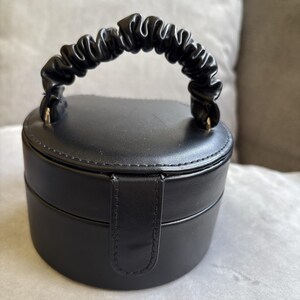 May include: A black, round jewelry box with a handle. The box is made of a smooth material and has a small clasp closure. The handle is a gathered, textured material. The jewelry box is sitting on a light gray surface.