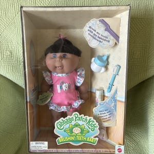 May include: A Cabbage Patch Kids Brushin' Teeth Baby doll in a box with accessories including a blue and white bottle, a toothbrush, and a tube of toothpaste. The doll has dark hair, blue eyes, and wears a pink dress with a tooth graphic.