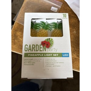 Pode incluir: Uma caixa branca de luzes LED em forma de abacaxi para uma festa no jardim. A caixa diz "GARDEN party PINEAPPLE LIGHT SET" e inclui 10 luzes brancas quentes. O comprimento iluminado é de 1,37 m.