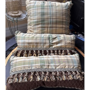 May include: Two decorative pillows with a plaid pattern in shades of green, beige, and brown. The rectangular pillow features a brown border with tassels and decorative trim. The square pillow is a matching plaid design.