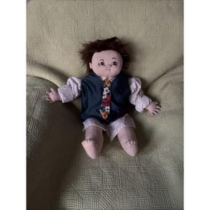 May include: A handmade doll with brown hair, wearing a white shirt, shorts, and a dark blue vest. The doll has a floral tie with red, yellow, and green accents. The doll is sitting on a green quilted surface.