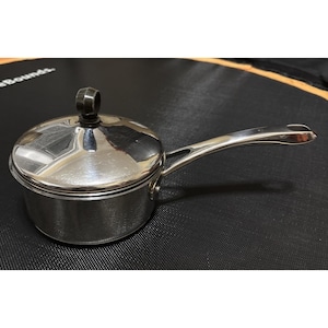 May include: A small, stainless steel saucepan with a lid and long handle. The pot has a shiny, reflective surface and a dark handle on the lid. The handle is curved for easy gripping. The saucepan is designed for cooking.