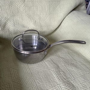 以下が含まれることがあります： 長いハンドルと透明なガラス蓋が付いたステンレス製の片手鍋。鍋の内側にはThomas Rosenthal Groupという文字が黒色で印字されています。鍋は光沢のある金属仕上げで、リベット留めされたハンドルが付いた曲線的な本体をしています。