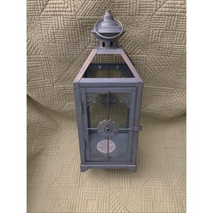May include: A rectangular metal lantern with a matte grey finish, featuring a hinged door with a circular filigree accent, decorative corner scrollwork, and a top handle. The lantern has clear glass panels and a small circular label on the base.