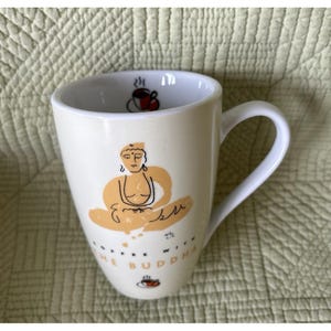 May include: A white ceramic mug featuring a stylized light orange illustration of a seated figure in a meditative pose. The text COFFEE WITH THE BUDDHA is printed in black below the figure, with small coffee cup icons above and below the design.