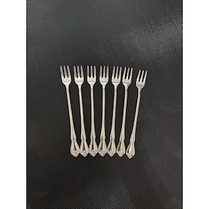 May include: A set of seven silver-toned metal cocktail forks, each featuring three tines and a decorative floral embossed pattern on the handle. These slender utensils are arranged in a parallel row, showcasing a polished finish and a classic design.