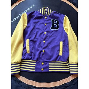 May include: A purple and yellow varsity jacket with the letter "B" on the chest. The jacket has yellow sleeves, a striped collar, and striped cuffs. The jacket is buttoned up and appears to be made of a satin-like material.