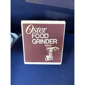 Peut inclure: Une boîte marron et blanche pour une tête de hachoir alimentaire Oster. La boîte présente le logo Oster en écriture blanche, avec les mots "FOOD GRINDER HEAD" en gras, en police sans-serif. Un dessin au trait de la tête du hachoir est en dessous.