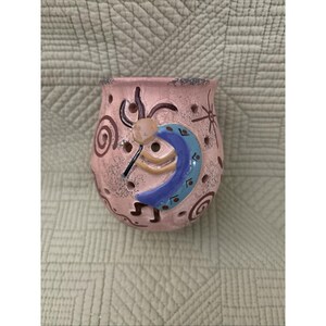 以下が含まれることがあります： ピンク色の陶器製アロマウォーマー。ココペリのデザインが施されています。青、黄、黒のココペリの姿、装飾的な穴、渦巻きと星の模様が特徴です。ウォーマーの高さは約10cmです。