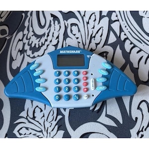 May include: A blue and gray Mathshark calculator with a digital display. The calculator has a variety of buttons for mathematical functions, including addition, subtraction, multiplication, and division. The calculator has the word "MATHSHARK" printed on the top.