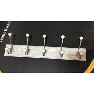May include: A rectangular wall-mounted coat rack featuring a distressed white finish with a beaded border. It includes five silver-toned metal hooks, each equipped with two white ceramic ball-tipped ends for hanging garments or accessories.