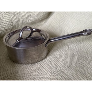 Puede incluir: Un cazo de acero inoxidable con tapa a juego y un mango largo y recto. La olla presenta un acabado metálico pulido y reflectante, un cuerpo redondeado y un asa curva en la tapa para facilitar su manipulación y levantamiento.