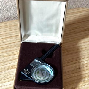 Könnte beinhalten: Ein schwarzer Kurvimeter mit einem runden Zifferblatt, das grüne und weiße Markierungen sowie das Wort MILES auf der Anzeige aufweist. Das Gerät befindet sich in einem beigen, rechteckigen Etui mit einer dunkelbraunen Samteinlage.