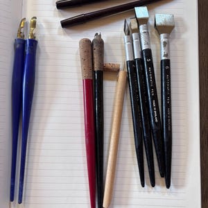 May include: An assortment of calligraphy pens and nibs are arranged on a lined notebook. The pens vary in color, including blue, red, black, and natural wood tones. Some pens have metal nibs, and others have cork handles. The notebook is open to a page with the months of the year.