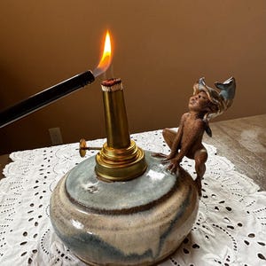 May include: A ceramic oil lamp with a brass burner and a small figurine. The lamp has a blue and cream glaze. A lighter is igniting the wick, producing a bright orange flame. The figurine is a small, brown figure with blue accents.