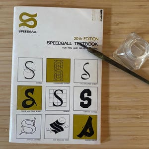 以下が含まれることがあります： ペンとブラシのレタリングのためのSpeedball 20th Edition教科書は、金色のロゴと様々なスタイルの9つの黒い文字Sの例が載った白い表紙が特徴です。暗い色の軸のカリグラフィーペンと透明なガラス製のインク壺が本の上に置かれています。
