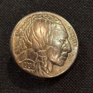 May include: A vintage 1936 Buffalo nickel coin. The coin features a detailed profile of a Native American on the obverse, with the word "LIBERTY" visible. The year "1936" is also displayed. The coin is a silver-colored metal.