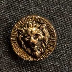 Pode incluir: Um botão redondo, de cor dourada antiga, com uma cabeça de leão detalhada no centro. A face do leão é rodeada por uma borda texturizada. O botão é provavelmente feito de metal e está sobre um fundo de tecido escuro.