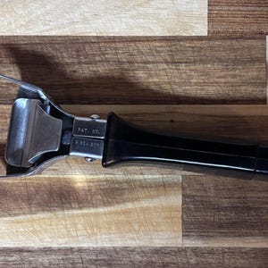 Può includere: Un manico per pentola nero e argento con un morsetto in metallo e un manico nero. Il manico ha un anello in metallo argentato all'estremità. Il manico è appoggiato su una superficie di legno.