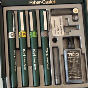 Pode incluir: Um conjunto de desenho Faber-Castell verde com quatro canetas TG1-S, um frasco de tinta de desenho Higgins e acessórios. As canetas têm diferentes tamanhos de ponta, incluindo 0,25 e 0,40. A tinta é preta mate.