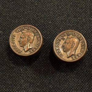 Puede incluir: Dos gemelos vintage de color cobre. Cada gemelo circular muestra el perfil de un hombre y la inscripción "GEORGIVS V D.G. BR: OMN: REX". Sobre un fondo oscuro y texturizado.