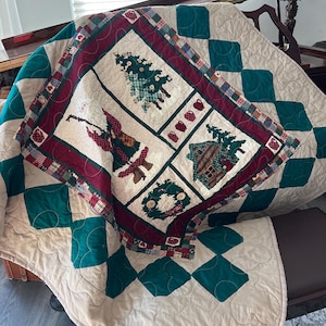 May include: A cream-colored quilted throw blanket with a green and red border. The border features a diamond pattern with a scalloped edge. The center of the blanket features a Christmas-themed design with a tree, a cabin, a snowman, and a wreath.