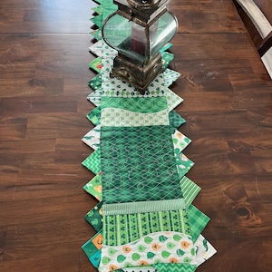 May include: A green and white table runner with a shamrock and clover pattern. The runner is made of fabric and has a scalloped edge. A lantern is sitting on top of the runner.