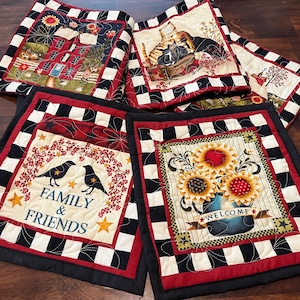 May include: A collection of quilted pot holders with various designs. One features a house, another a cat, and a third displays sunflowers in a vase with the word "WELCOME". Each has a black and white checkered border and red accents.