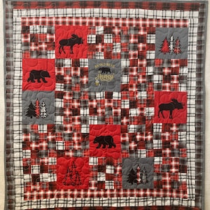 May include: A red and black plaid quilt with a patchwork design. The quilt features embroidered images of bears, moose, and pine trees. The text "An old bear and his honey live here" is embroidered in the center of the quilt.