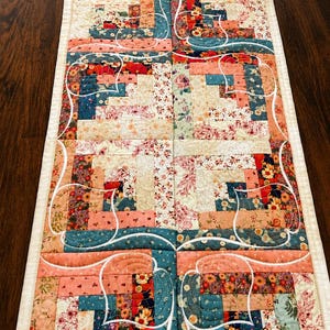 May include: A floral patchwork table runner, measuring 13" x 25", features a mix of floral and geometric patterns in shades of pink, teal, and cream. The runner is bordered with a white edge and has decorative white accents.