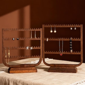 May include: Two wooden jewelry organizers with a dark brown finish. Each organizer has multiple rows of small holes for hanging earrings and necklaces. Various earrings and necklaces are displayed on the organizers. The organizers sit on a light-colored surface.