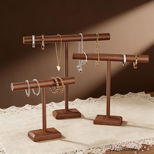 May include: Three wooden jewelry display stands in varying heights. Each stand has a horizontal bar for hanging necklaces, bracelets, and rings. The stands are made of dark brown wood and are displayed on a cream-colored surface.