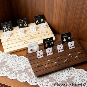 May include: Two tiered wooden jewelry display stands with horizontal grooves for rings and vertical slots for earring cards. One stand is light natural wood, the other is dark brown wood. Both feature various earrings and rings on display.