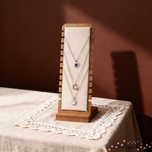 May include: A wooden jewelry display stand with three silver necklaces, each featuring a different pendant: a blue heart, a circle, and a pink star. The stand is on a cream-colored surface, with a brown background.
